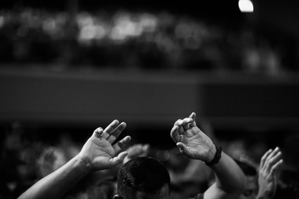 grayscale-of-photo-of-man-2014774 A black and white photo of hands raised in a crowd during a concert or religious gathering indoors.